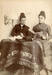 portrait en buste de deux femmes assises derrière une table couverte d'une nappe sur laquelle est posée une lampe, une tenant un livre